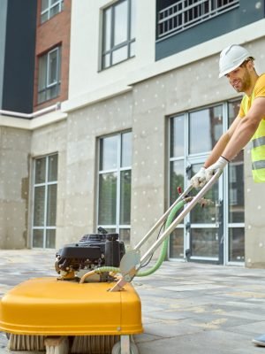 Working process. Smiling worker in protective helmet gloves and yellow tshirt cleaning construction site using special equipment and two men discussing looking at new building