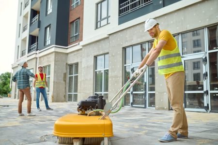 Working process. Smiling worker in protective helmet gloves and yellow tshirt cleaning construction site using special equipment and two men discussing looking at new building